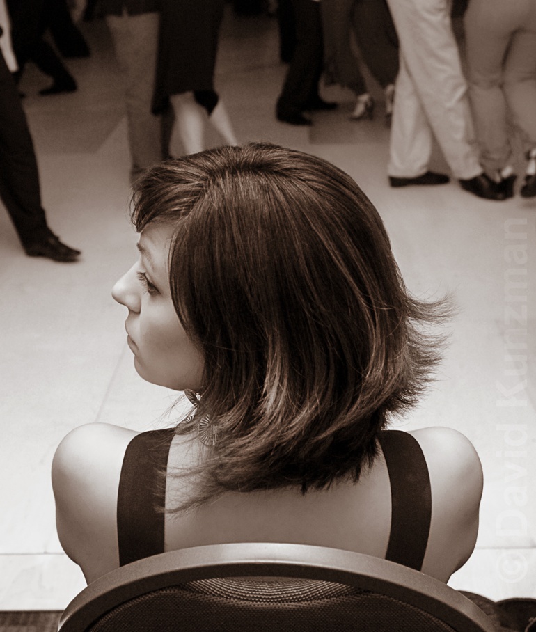 A photograph of my friend taking a break from dancing during a milonga.  She looks to the side as I take her photo from behind.  Tango dancers are dancing in the background.