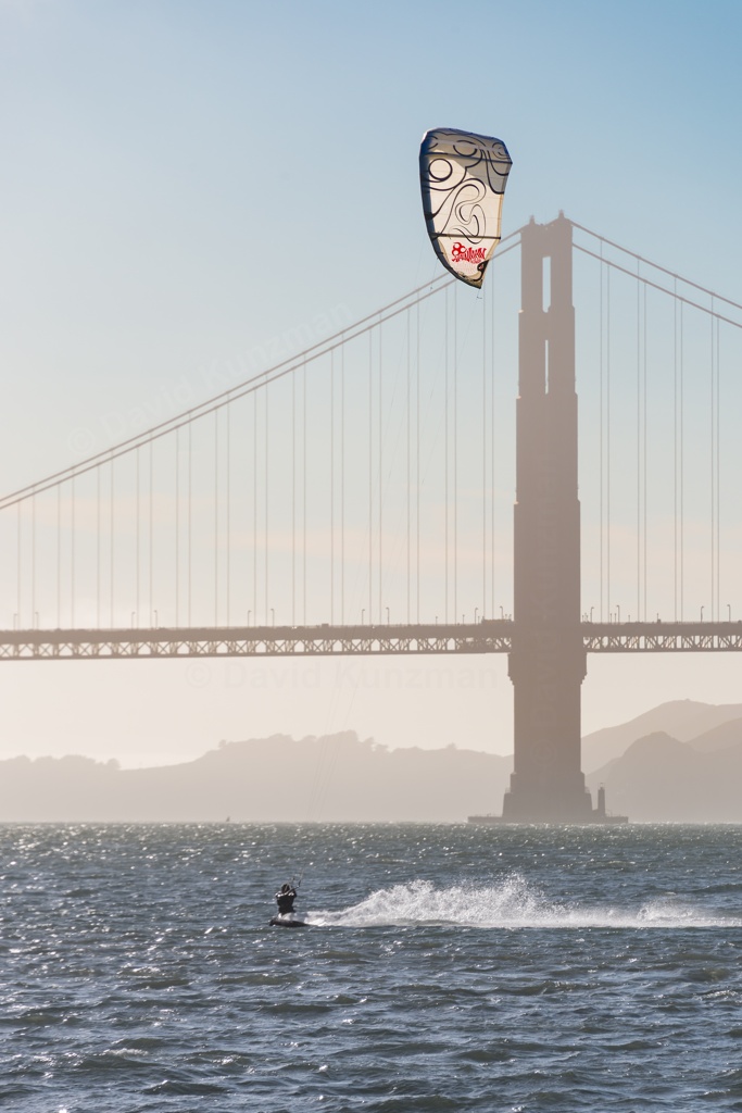 A kite boarder passing in front of one of the Golden Gate Bridge's towers, under clear skys and with the rolling hills of the Marin Headlands in the background.