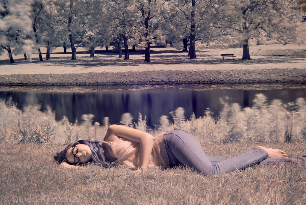An infrared photograph of Mandy lying down in a grass field in front of a pond.  The false color image shows the plants as white and her hair as blue.