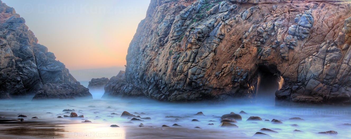 A long exposure photograph of waves flowing between rocks on Pfeiffer Beach, CA as the sun sets in the background.