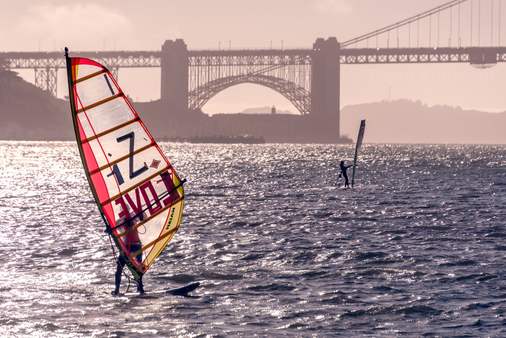 A couple windsurfers out on the water with the Golden Gate Bridge in the background, one with "Zi Love" written across their sail.