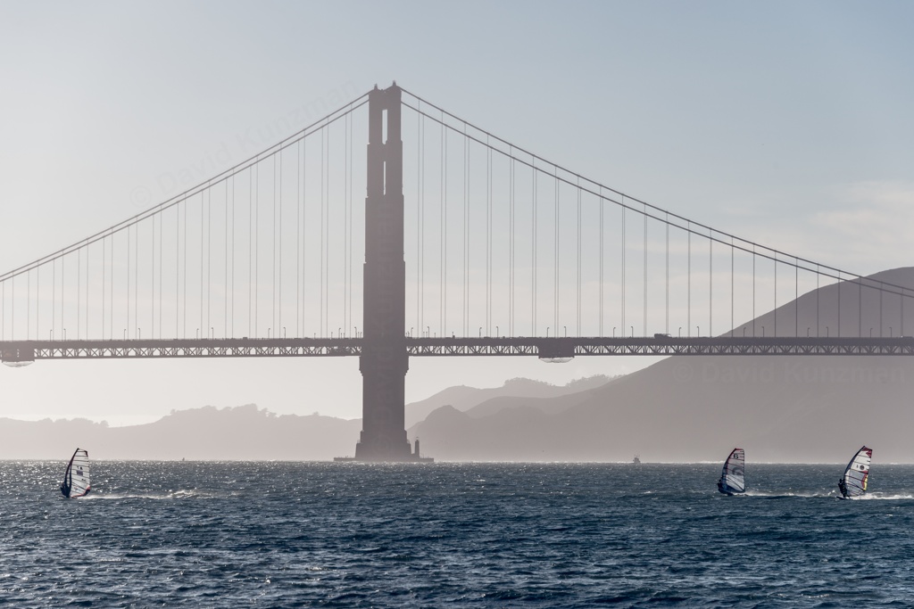 Three windsurfers passing in front of one of the towers of the Golden Gate Bridge, with clear skys above and rolling hills of the Marin Headlands in the distant background.